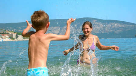 Cheerful laughing mother with little boy playing and splashing water at the sea beach. Family holiday, vacation and fun summertime of children and parentsの写真素材