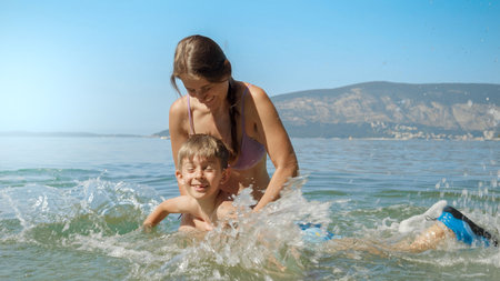 Happy cheerful boy splashing sea water with feet while learning swimming on the beach with mother. Family holiday, vacation and fun summertime of children and parentsの写真素材