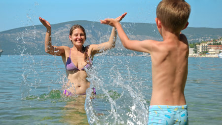 Happy laughing mother with son splashing sea water and having fun on beach. Family holiday, vacation and fun summertime of children and parentsの写真素材