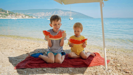 Two adorable boys enjoy sweet watermelon on the sea beach. Essence of a summertime getaway, holiday merriment, and the simple pleasures of vacation.の写真素材