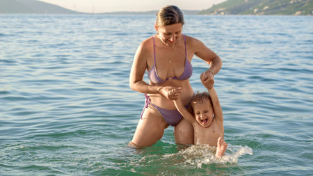 Young smiling woman in bikini holding her baby son by hands while having fun in the sea. Summertime, holiday, vacation, happiness and relaxationの写真素材