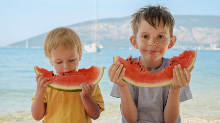 Two cute boys savoring sweet watermelon on a sea beach towel. Spirit of summertime, holiday delights, vacation happiness, and the relaxation of sunny days.の写真素材