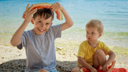 Two funny smiling boys enjoying eating watermelon and playing with rinds on the sea beach. Summertime, holiday, vacation, happiness and relaxationの写真素材