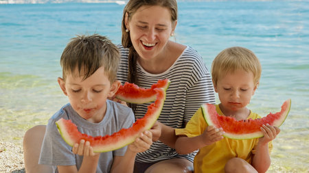 Mother and her two boys enjoy sweet watermelon on a beachside picnic. Family bonding during a holiday, summertime merriment, and the simple pleasures of a vacation.の写真素材