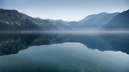High mountains reflecting in calm sea waves with rising morning fog or mist.