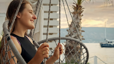 Portrait of beautiful smiling woman enjoying swinging and looking on the sunset sky over the sea beach. Concept of summertime, vacation and beach holidaysの写真素材