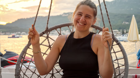 Portrait of smiling woman relaxing in rope eat and swinging against sunset sky over sea beach.の写真素材