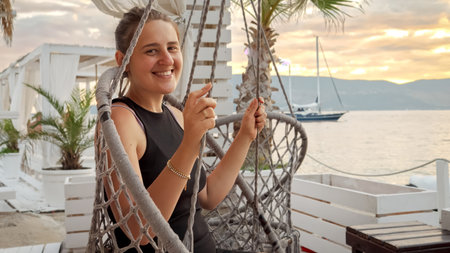 Cheerful smiling woman having fun on swing at sea beach bar at sunset.の写真素材