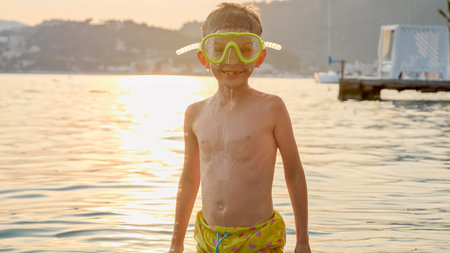Happy cheerful boy in diving mask for snorkeling standing in a calm sea waves against sunset light rays.の写真素材