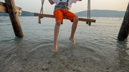 Closeup of little boy swinging on the sea beach and touching ocean waves with feet. Holiday, summer vacation and tourism.の写真素材