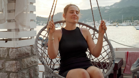 Young smiling woman relaxing and swinging in rope nest or chair swing in sea beach bar at the sunset. Concept of holiday, summertime, relaxation in resort, vacation, sea trip and journeyの写真素材