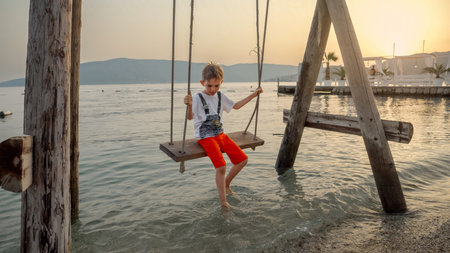 Little boy swinging on wooden rope swing on the sea beach at sunset. Holiday, summer vacation and tourism.の写真素材