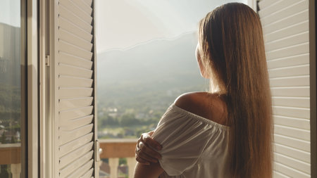 Young brunette, dressed elegantly, opens her window blinds, ventures to the terrace, and looks at...