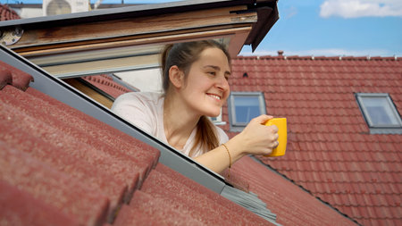 Happy smiling woman opens attic window and drinks tea or coffee in the morning. She is enjoying city...