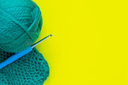 A metal hook with a blue handle, a skein of green yarn and a part of a knitted fragment lies on the side of the photo on a yellow background. top view, flat lay, copy space, isolateの写真素材