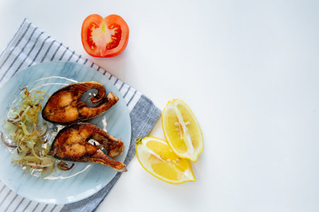 Fried fish with onions lies on a plate, on a waffle towel, next to a lemon and a tomato on a white background.の写真素材