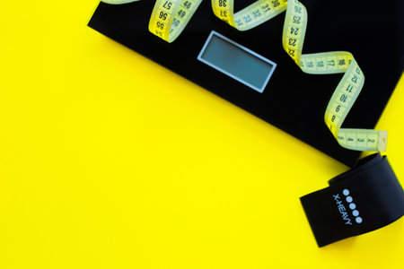 A weight scale, a black fitness tape and a yellow measuring tape lie on a yellow background on the side of the photo. top view, flat lay, copy space, isolate.の写真素材
