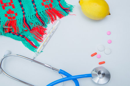 A warm winter scarf, a thermometer, a lemon, a stethoscope and pills with capsules lie on a white...