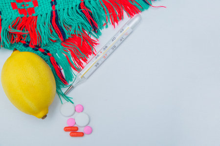 A warm winter scarf, thermometer, lemon and pills with capsules lie on the side on a white...