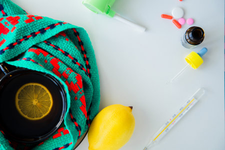 A cup of tea with lemon stands on a knitted green and red scarf, next to it is a thermometer, lemon...