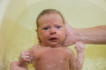 A newborn baby is bathing in a bath for the first time, his hand is holding his head, the baby is...