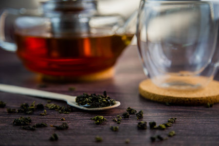 A wooden spoon with tea lies against a dark wood background, a mug and a teapot are visible from...