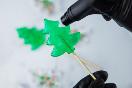 A green lollipop in the form of a Christmas tree is held in the hands of black gloves and burned on fire from a lighter, in the process of making a lollipop. High quality photoの写真素材