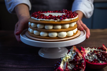 The cake stands on a white stand, decorated with cream and pomegranate on top, it is held by the hands of a pastry chef, there is a pomegranate next to it and there are flowers on a dark background. High quality photoの写真素材