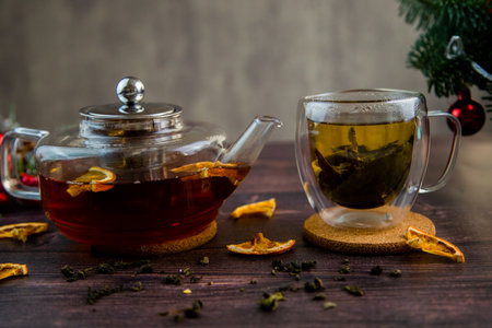 A transparent teapot and a cup of tea stand against a background of dark wood, slices of dried orange and Christmas decorations. High quality photoの写真素材