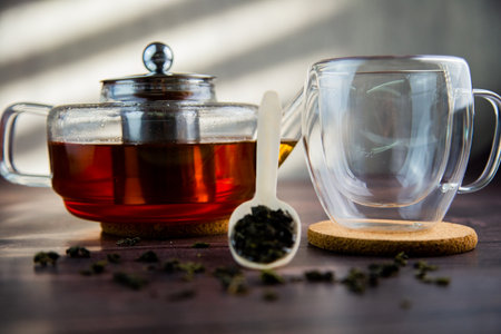 A wooden spoon with tea lies against a dark wood background, a mug and a teapot are visible from behind, a photo in dark colors. High quality photoの写真素材