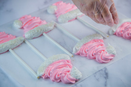 A pastry chefs hand in a transparent glove sprinkle coconut shavings on a pink meringue cap on a...