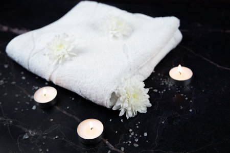 A white towel lies on a dark background surrounded by white flowers and burning candles. High...