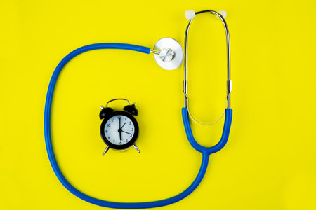 Black alarm clock lies inside a blue phonendoscope on a bright yellow background. High quality photoの写真素材