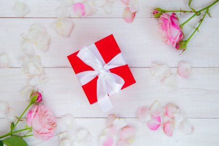 On a white wooden background there are roses and petals forming a circle, inside there is a gift in...