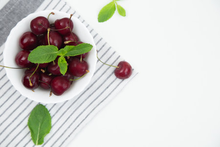 A white plate with a ripe burgundy cherry and a mint petal stands on a striped kitchen towel on a white background with a place for text, top view. High quality photoの写真素材