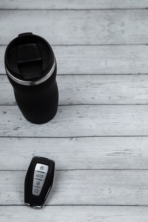 A car key with automatic buttons and a thermocup are black with metal inserts on a white wood...