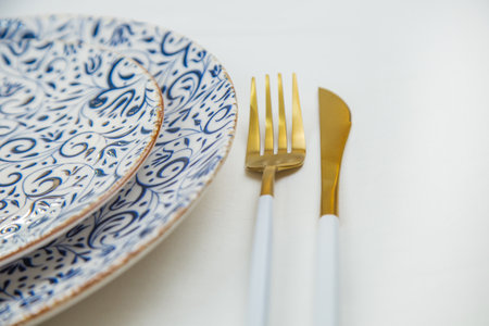 On the side, two serving plates with blue patterns on a white background stand one on one, part of the plates is visible, gold-white appliances are located on the side, a close-up side view on a white background. The concept of serving. The concept of eating. High quality photoの写真素材
