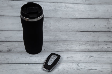 A car key with automatic buttons and a thermocup are black with metal inserts on a white wood background with a place for text. High quality photoの写真素材