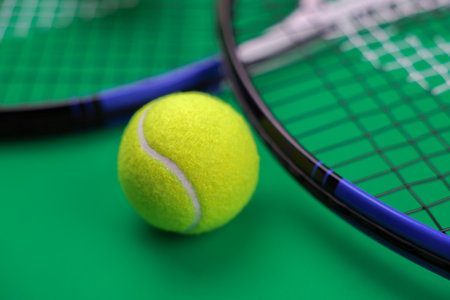 A yellow lawn tennis ball in close-up lies near two lawn tennis rackets on a green background. The...