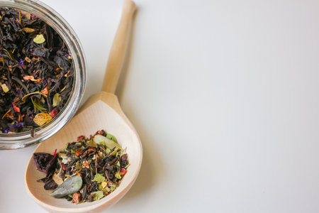 Large-grain tea with the addition of pieces of fruit and berries lies on a wooden spoon, next to it is a tilted glass jar with tea on a white background with space for text, Concept of tea drinking. High quality photoの写真素材