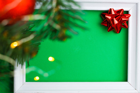 A white wooden frame is located in the center of the photo on a green background, from the left side you can see green branches of a Christmas tree, a golden garland and a red shiny ball, on the right of the frame is a red bow. Concept of Holiday, New Year and Merry Christmas. High quality photoの写真素材