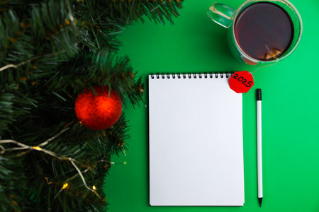 A white pencil with a black lead and a black eraser lie on a green background, next to it is a mug of black tea with a paper red label wishing a Happy New Year, on the left there are branches of a Christmas tree with a red ball and a yellow garland. The concept of a holiday, a New Years mood. High quality photoの写真素材