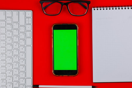 A phone with a green chroma key screen is lying on a red background, to the right of it is a white notebook with black springs on top, black glasses with transparent glasses are on top, a white computer keyboard is on the left, and a white pencil with a black eraser and a black lead is at the bottom. High quality photoの写真素材