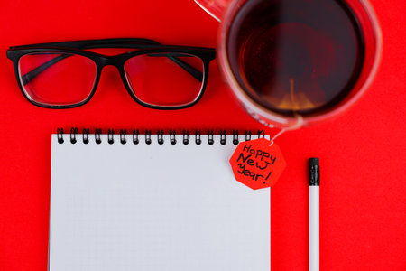 A white notebook with a black spring at the top lies in the center of the photo, next to it is a white pencil with a black eraser, transparent black-framed glasses and a mug of tea are on top, a red label stretches from the tea bag, where it is written, Happy New Year. High quality photoの写真素材