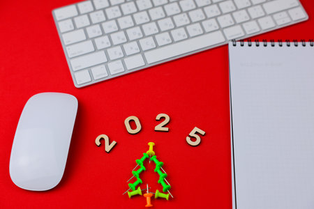 On a bright red background, there is a creative Christmas tree made of office nails, above it are wooden numbers 2025, a white computer mouse and keyboard are lying around, a white notebook with a black spring. The concept of an office, a holiday. High quality photoの写真素材