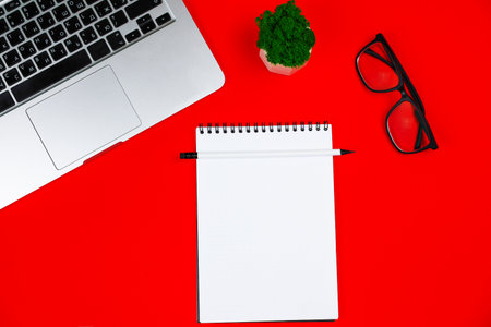 A white notebook with a black spring and a white pencil are lying on a red background, next to it are a gray laptop with a black keyboard, a green plant in a white pot: black-framed glasses and a red festive bow. Office and holiday concept. High quality photoの写真素材