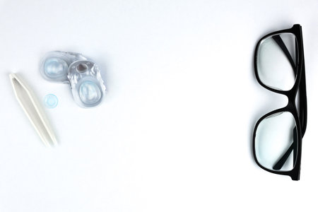 On the right of the photo there are black-framed glasses, on the left there are disposable containers with contact lenses, a blue lens and beige tweezers in close-up on a white background with space for text in the center. Vision, correction and ophthalmology concept. High quality photoの写真素材