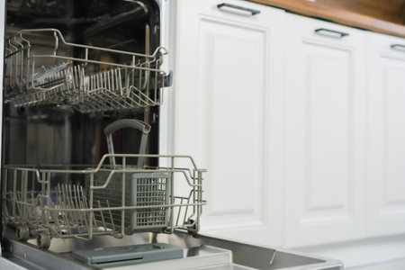 An empty dishwasher with two compartments in gray tones stands with an open door ready to be loaded to the left, to the right of it are white kitchen cabinets with a black handle and a wooden countertop. High quality photoの写真素材