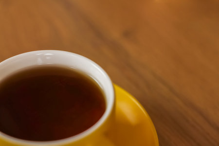 A yellow mug of black tea sits on a saucer in the lower left corner of the photo on a wooden...