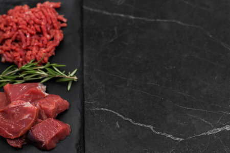 Top left view of chunks of beef lying on a black cutting board, separated by sprigs of rosemary with...